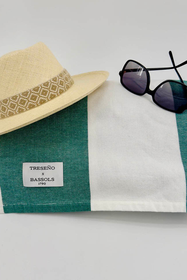A folded green and white striped linen scarf with a white TRESENO x BASSOLS 2023 label, styled with a beige straw hat and black sunglasses on a light gray background.