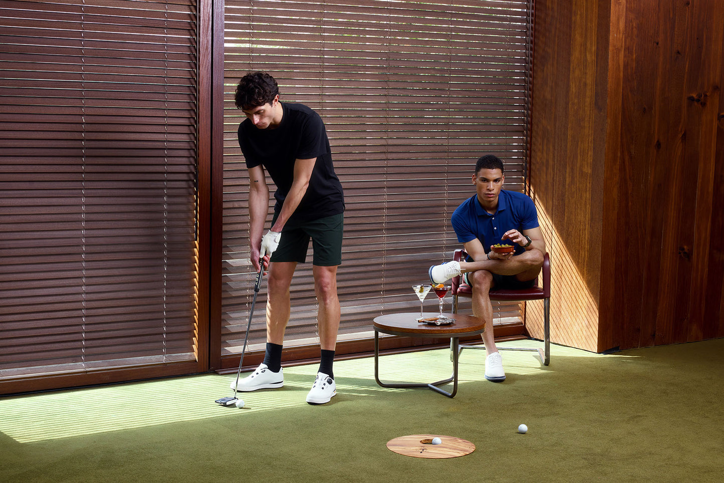 Two men playing practicing putting with a wooden wall and green floor.