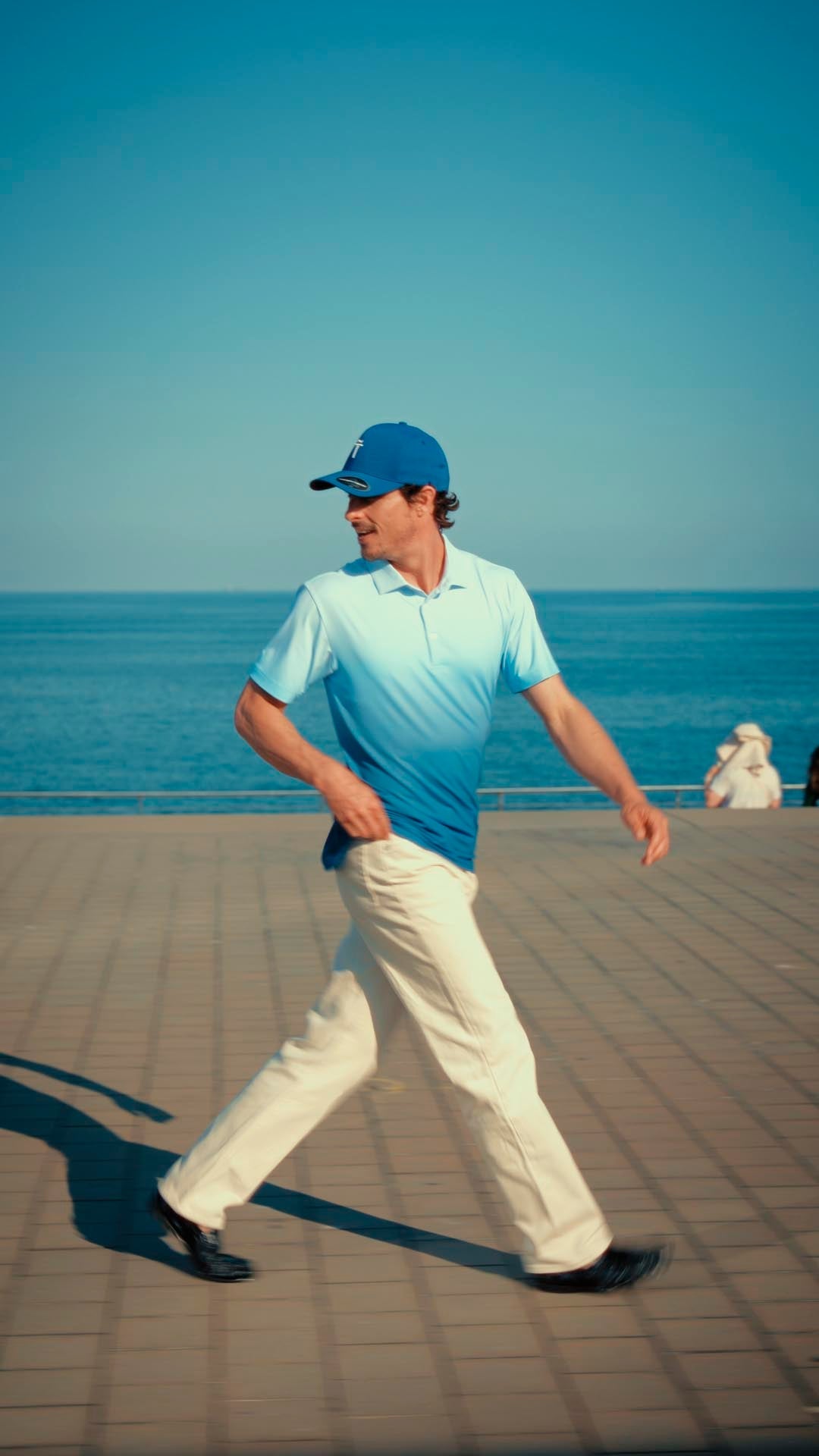 Man walking on a boardwalk by the ocean wearing a blue cap and light blue shirt.