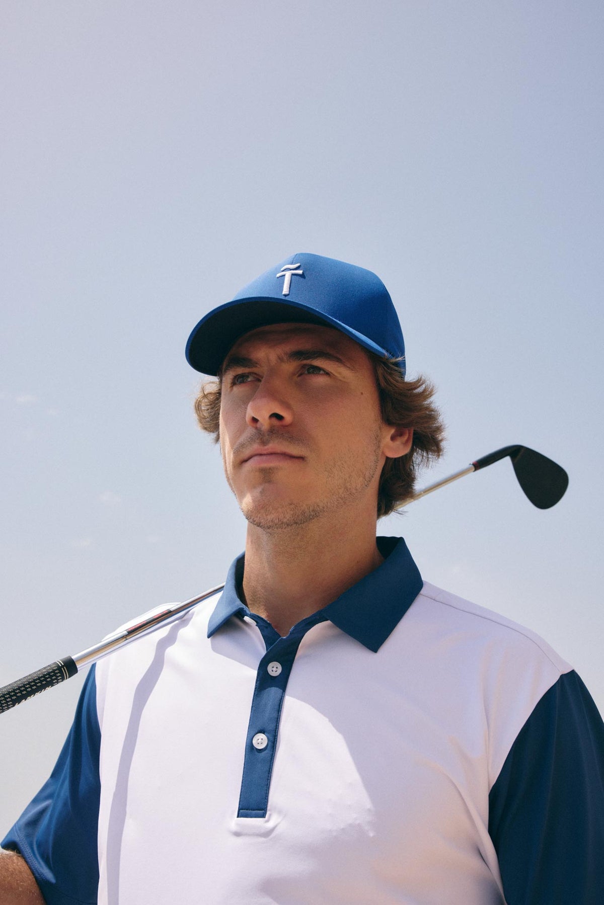 A man wearing a blue and white polo shirt and a blue cap with a white cross logo, holding a golf club on a sunny outdoor day.