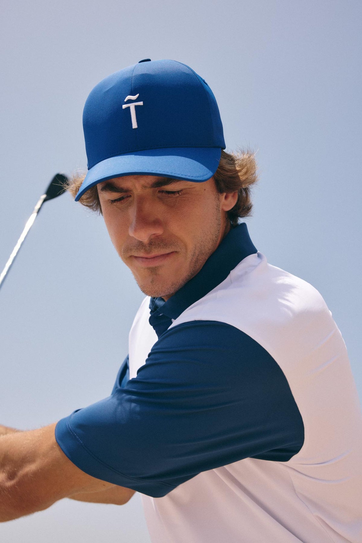 A man wearing a blue and white polo shirt and a blue cap with a white logo, captured mid-swing against a clear light blue sky background.