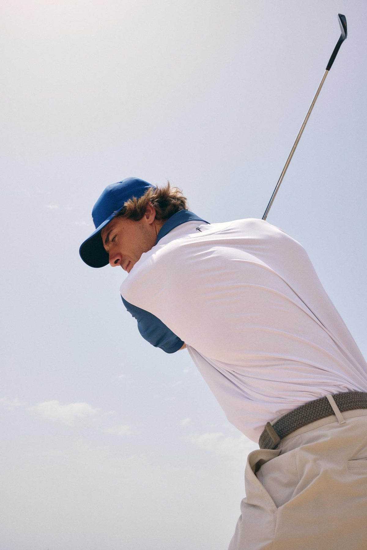 A golfer in a white polo shirt, beige pants, and a blue cap mid-swing against a bright sky background.