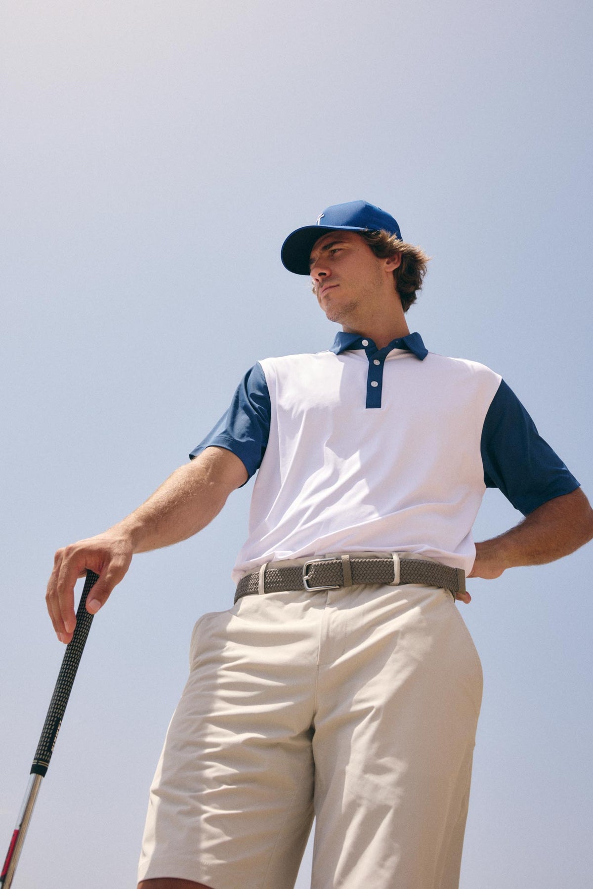 A man wearing a white and navy blue polo shirt, beige golf shorts, a blue cap, and a belt, holding a golf club against a clear blue sky.