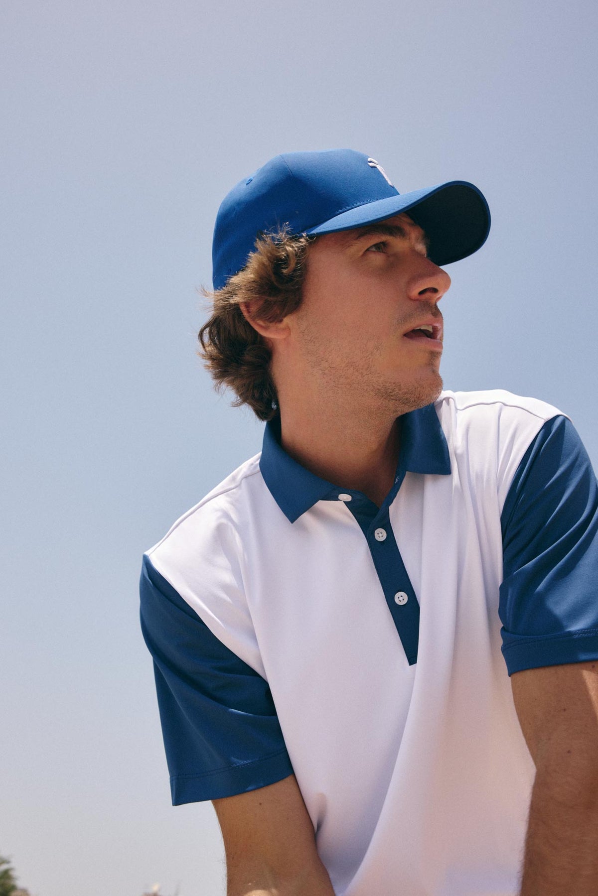 A man wearing a blue and white color-blocked polo shirt and matching blue cap, photographed outdoors against a clear light blue sky.
