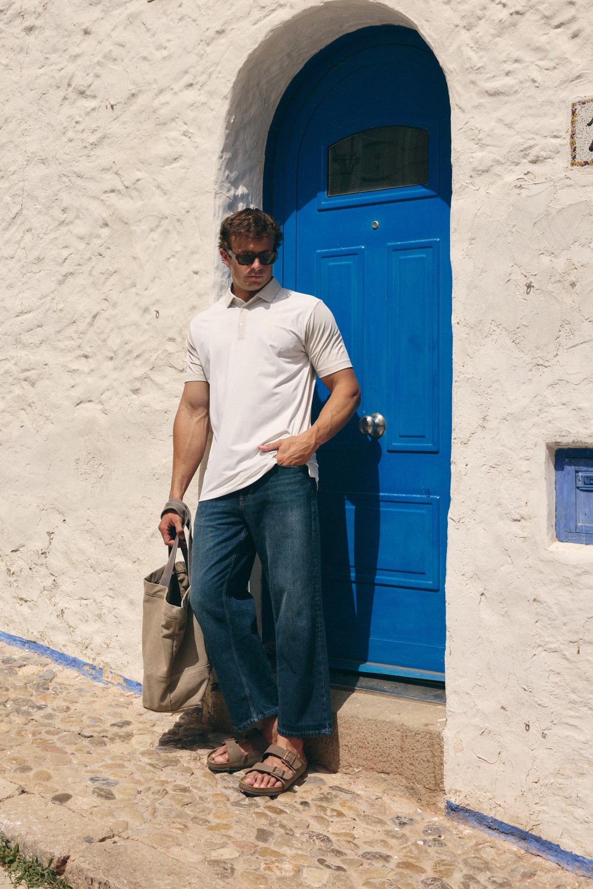 A man wearing a white short-sleeve polo shirt, dark blue jeans, and brown sandals stands in front of a white stucco wall with a bright blue door, holding a beige tote bag.
