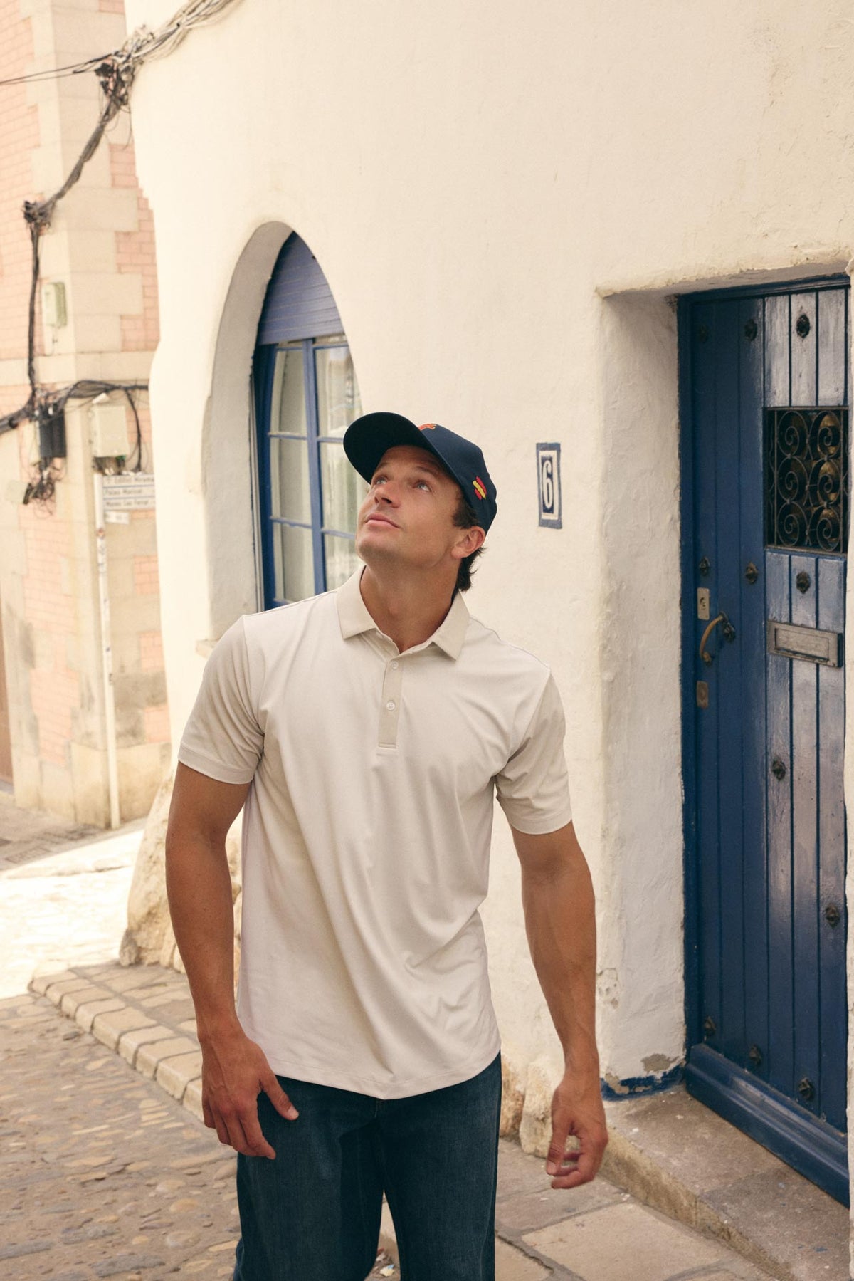 A man wearing a light beige short-sleeve polo shirt and a dark navy baseball cap stands on a cobblestone street in front of a white stucco building with blue doors.