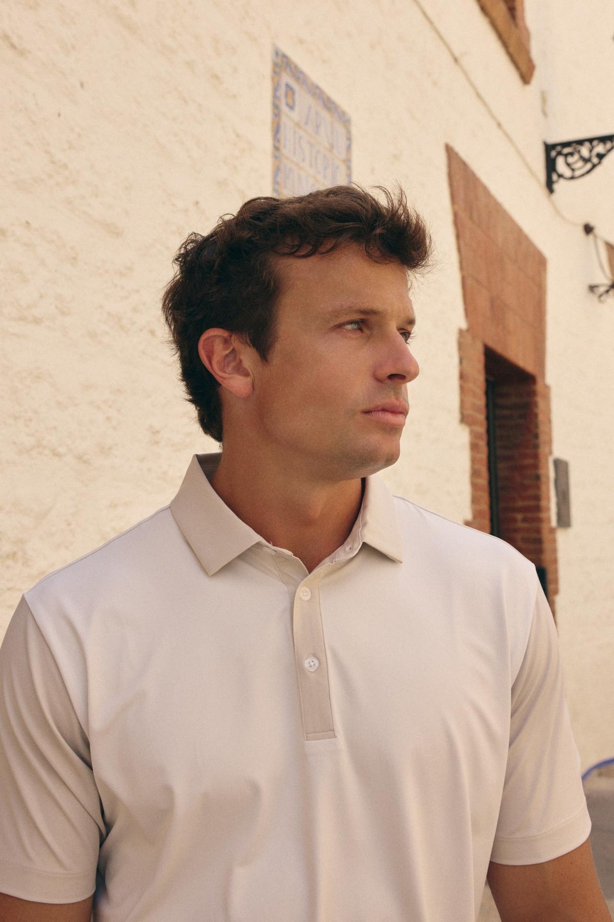 A man wearing a light beige short-sleeve polo shirt, standing outdoors against a textured white stucco wall.