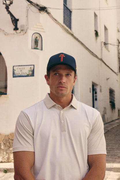 A man wearing a light beige polo shirt and a navy blue baseball cap with a red logo, standing in front of a white building with a street sign that reads 'Carrer de San Pablo'.