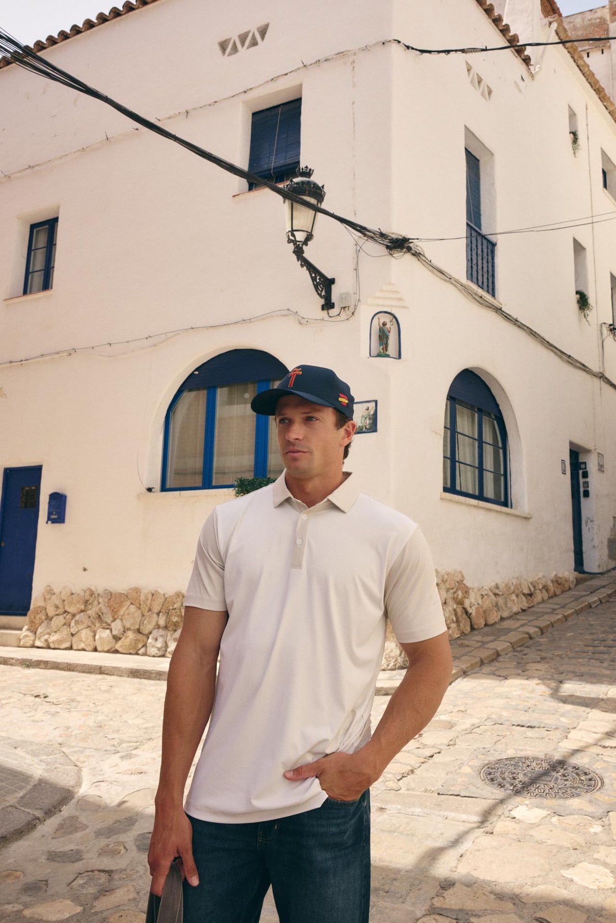 A man wearing a white short-sleeve polo shirt and a navy blue baseball cap stands on a cobblestone street in front of a white stucco building with blue-trimmed windows.