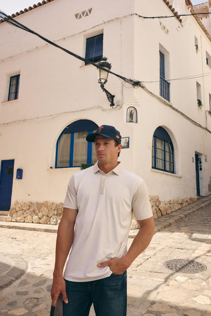 A man wearing a white short-sleeve polo shirt and a navy blue baseball cap stands on a cobblestone street in front of a white stucco building with blue-trimmed windows.
