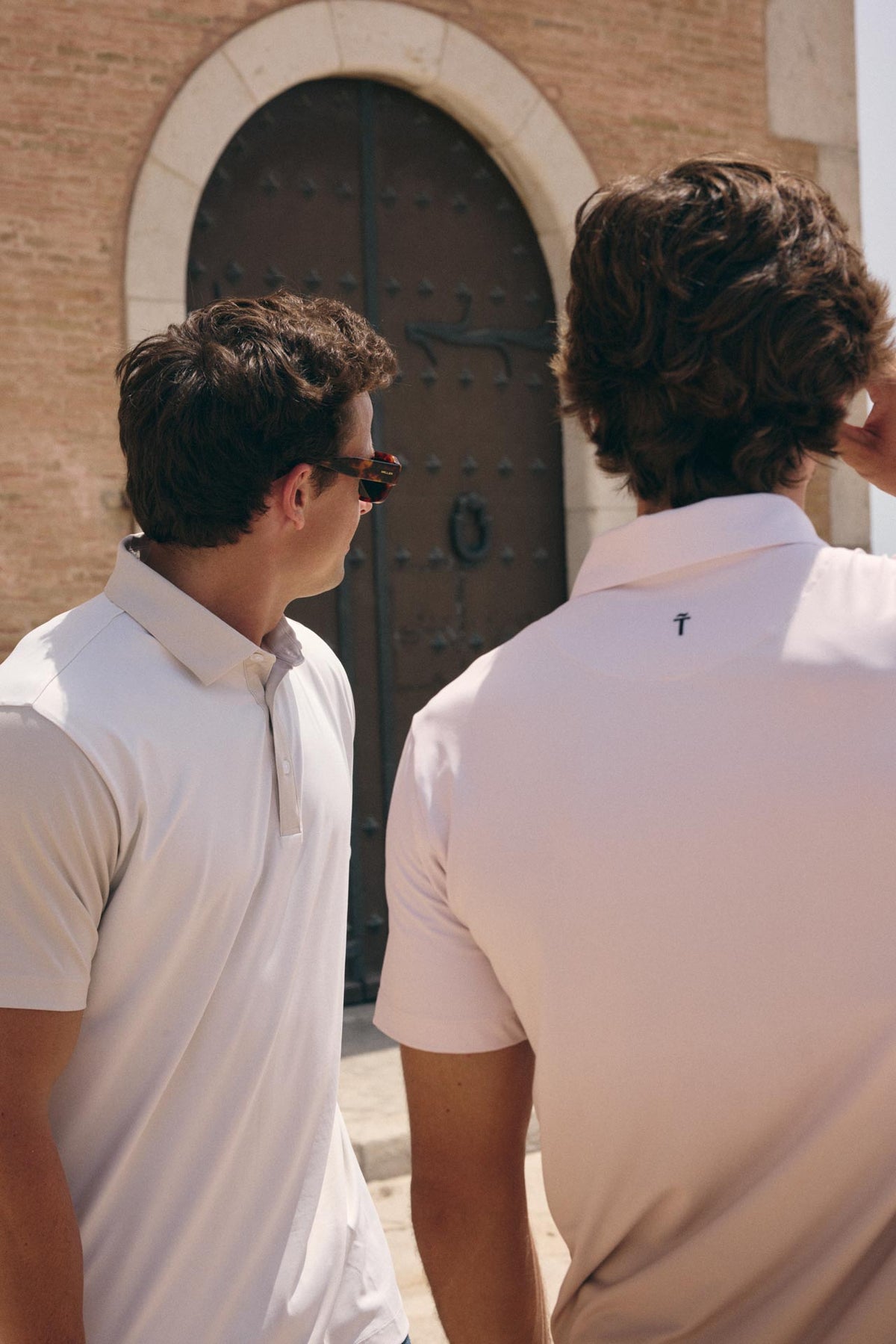 Two men wearing white polo shirts standing outdoors in front of a stone archway and dark wooden door. One man is shown from the back, revealing a small dark cross logo on the upper center of the shirt, and the other is shown in profile wearing sunglasses.