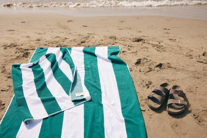 A green and white striped beach towel is laid out on sandy beach next to a pair of brown slide sandals, with ocean waves in the background.