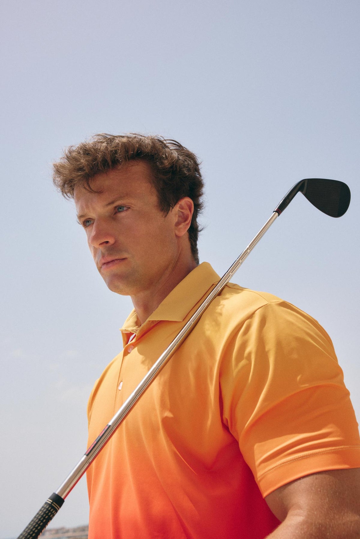 A man wearing a bright orange polo shirt and holding a golf club on a sunny outdoor day under a clear blue sky.