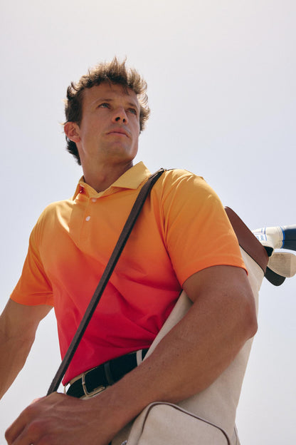 A man wearing a bright orange-to-red gradient polo shirt and beige trousers, carrying a golf bag over his shoulder against a pale sky background.