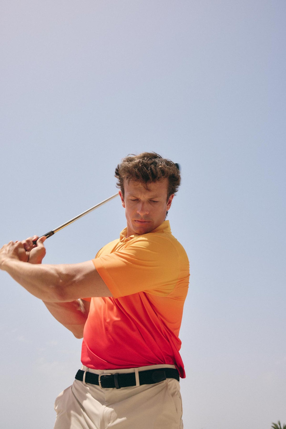 A man in a yellow-to-red gradient polo shirt and khaki pants swings a golf club against a clear blue sky.