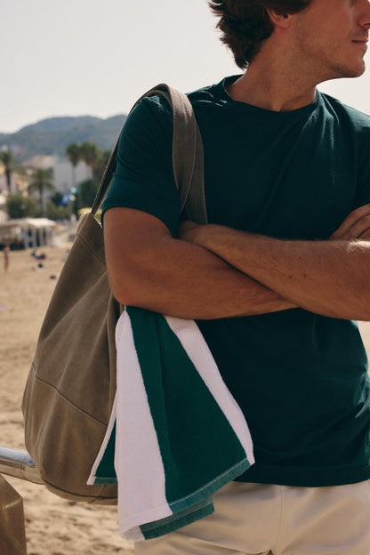 A man on a beach wearing a dark green t-shirt and light beige shorts, carrying a large tan shoulder bag and holding a matching green and white towel.