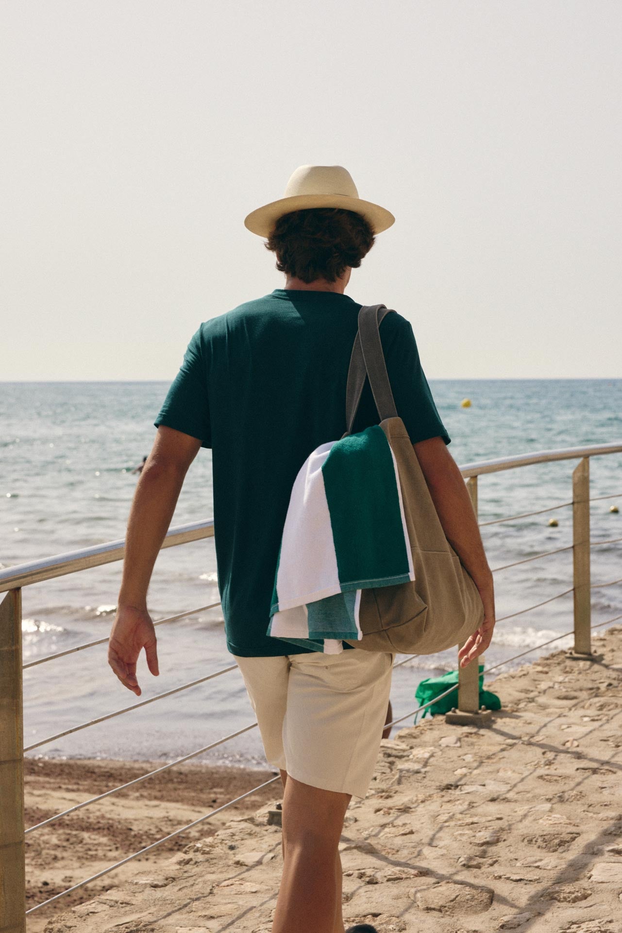 A man on a sandy beach walks away from the camera wearing a dark green t-shirt, beige shorts, and a cream-colored hat. He is carrying a large beige tote bag over his shoulder, which is folded with a green and white towel inside.