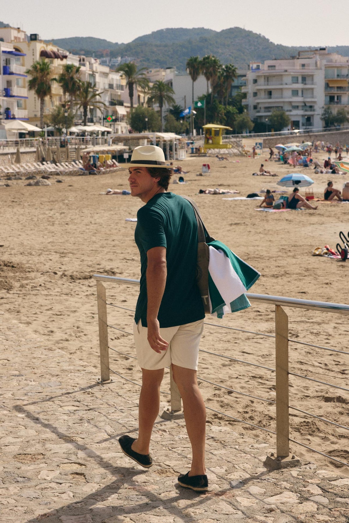 A man wearing a green t-shirt, cream shorts, and a straw hat walks along a sandy boardwalk at a beach, carrying a green and white shoulder bag.