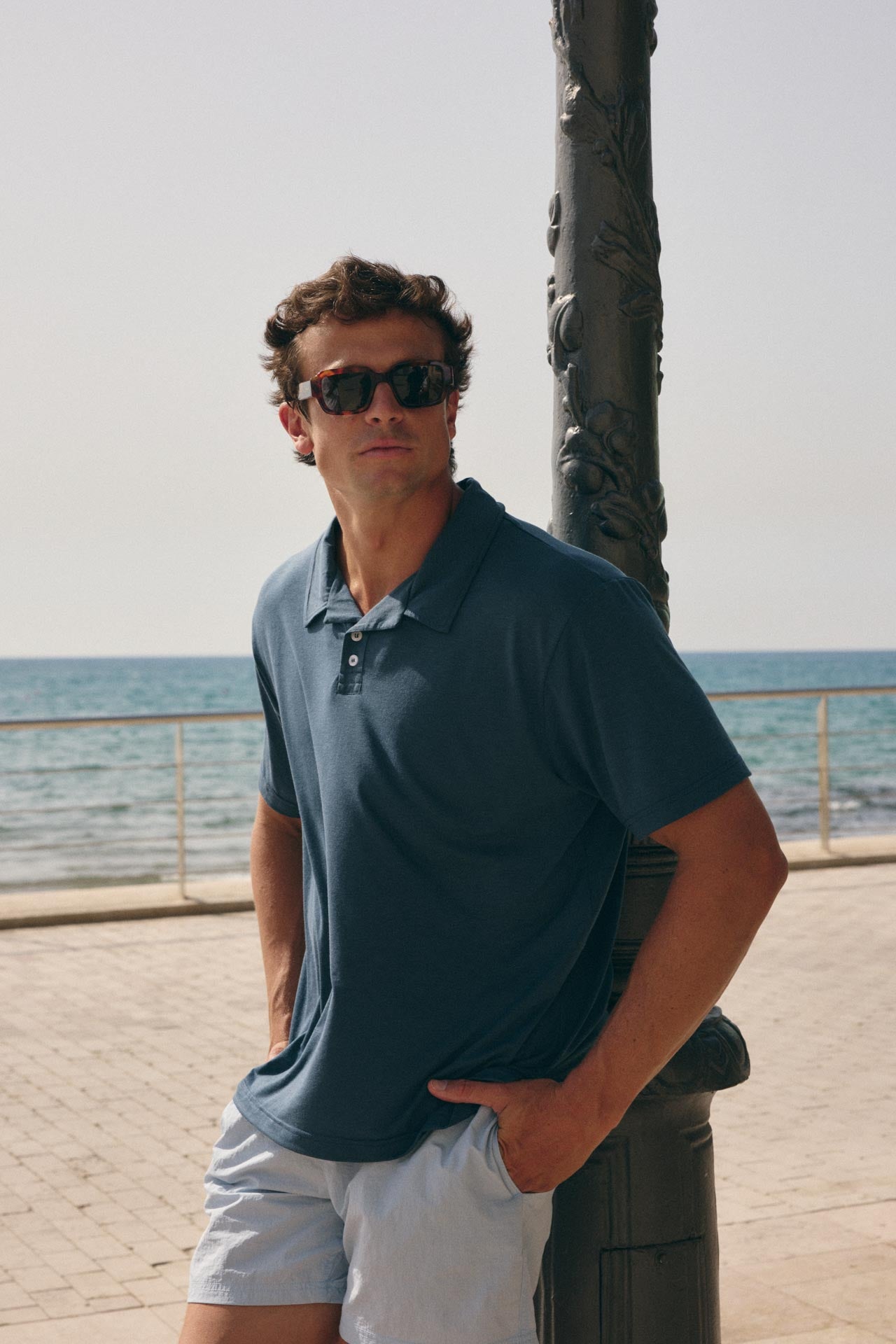A man wearing a navy blue polo shirt and white shorts stands outdoors by a seaside promenade, leaning against a dark metal pole and wearing dark sunglasses.