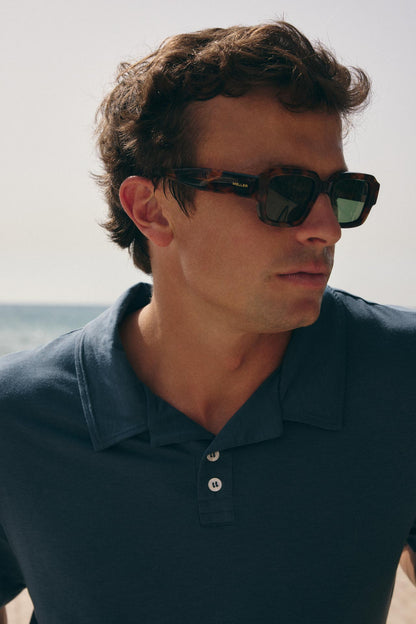 A man wearing dark tortoiseshell sunglasses with green-tinted lenses and a navy blue polo shirt, photographed outdoors against a bright sky and sea background.