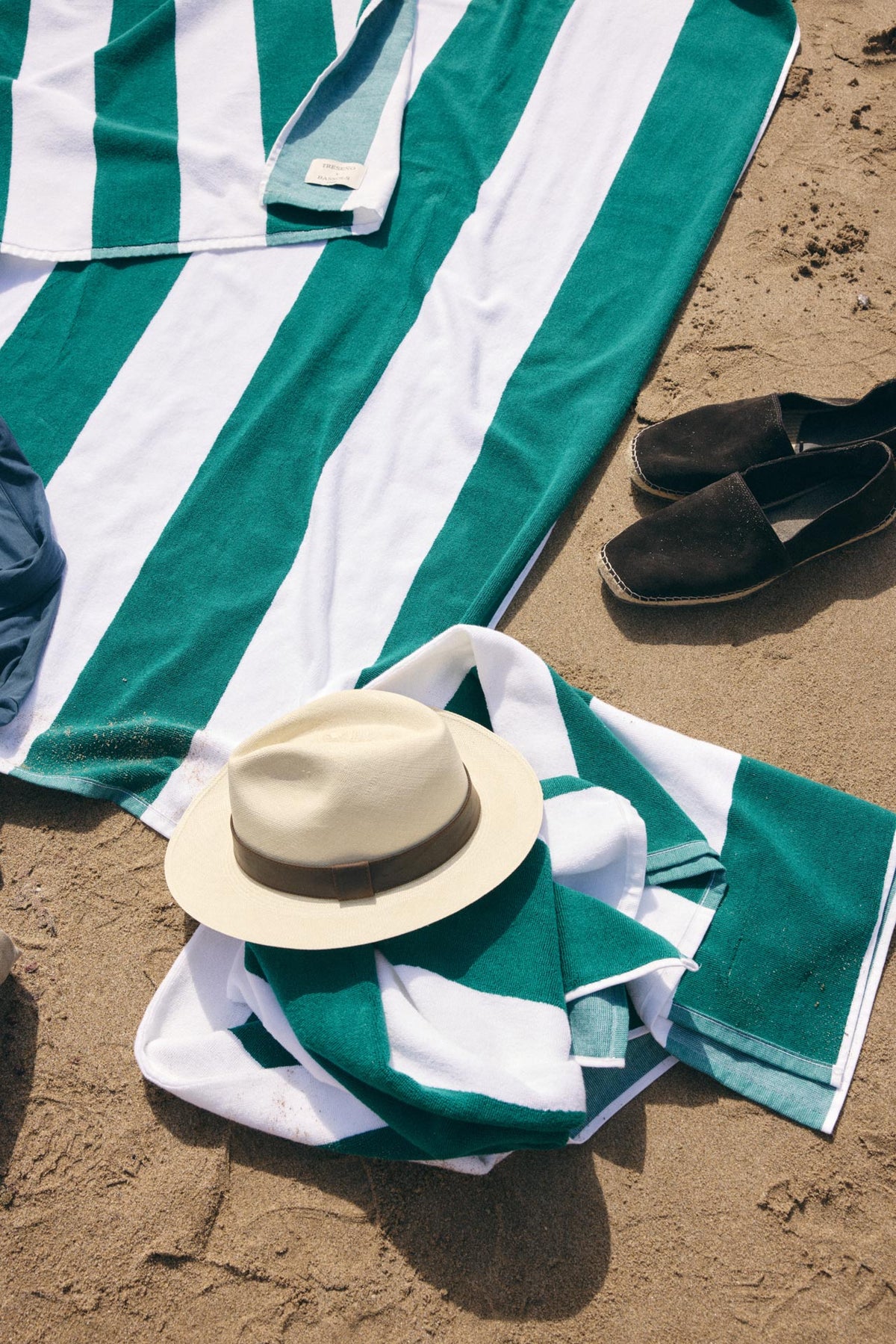 A green and white striped beach towel spread on sandy ground, styled with a luxury Panama hat, a matching folded towel, a pair of dark slip-on shoes