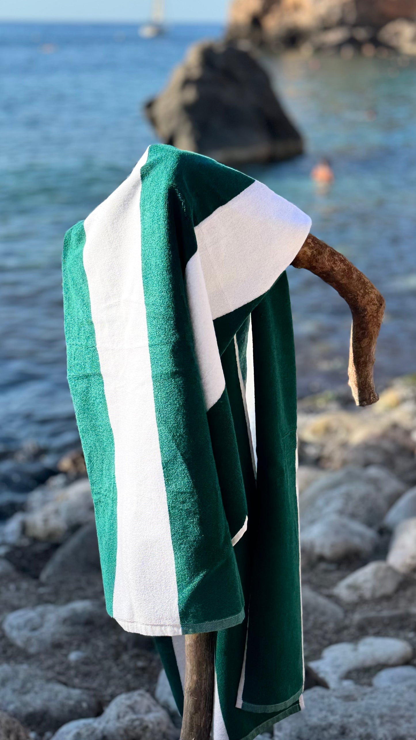 A green and white striped beach towel draped over a rustic wooden stand on a rocky shoreline with the sea in the background.