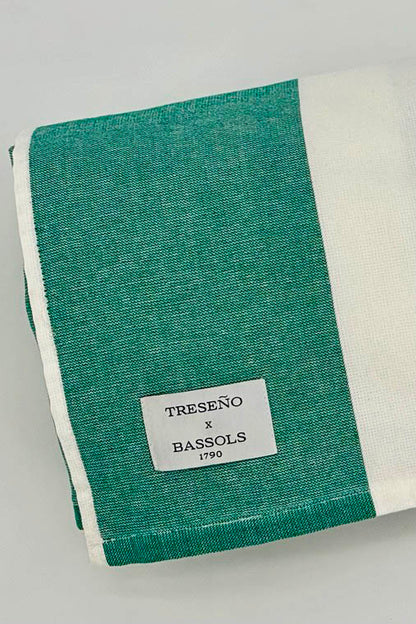 A folded striped textile towel or blanket in teal green and white, with a small sewn label reading TRESEÑO x BASSOLS 1990.