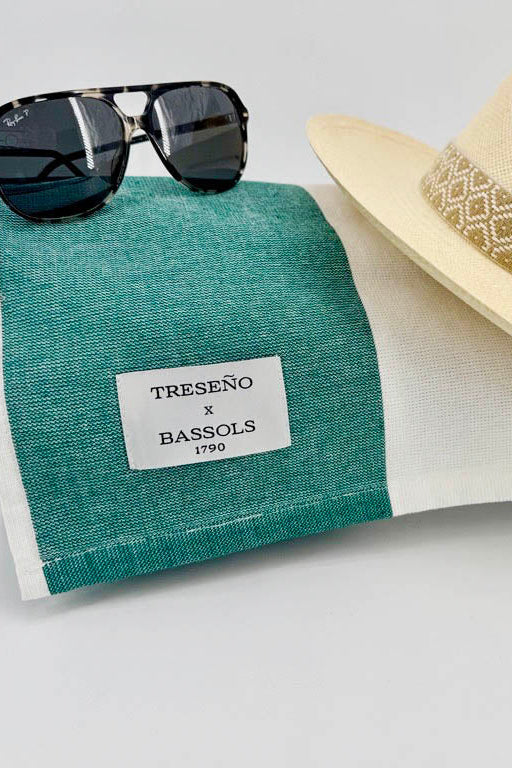 A folded teal and white striped linen sun towel with a white label reading 'TRESEÑO x BASSOLS 1790', styled with black sunglasses and a beige straw fedora hat on a light gray background.