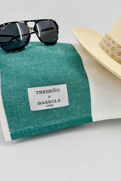 A folded teal and white striped linen sun towel with a white label reading 'TRESEÑO x BASSOLS 1790', styled with black sunglasses and a beige straw fedora hat on a light gray background.