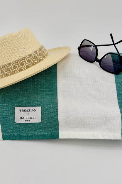 A folded green and white striped linen scarf with a white TRESENO x BASSOLS 2023 label, styled with a beige straw hat and black sunglasses on a light gray background.