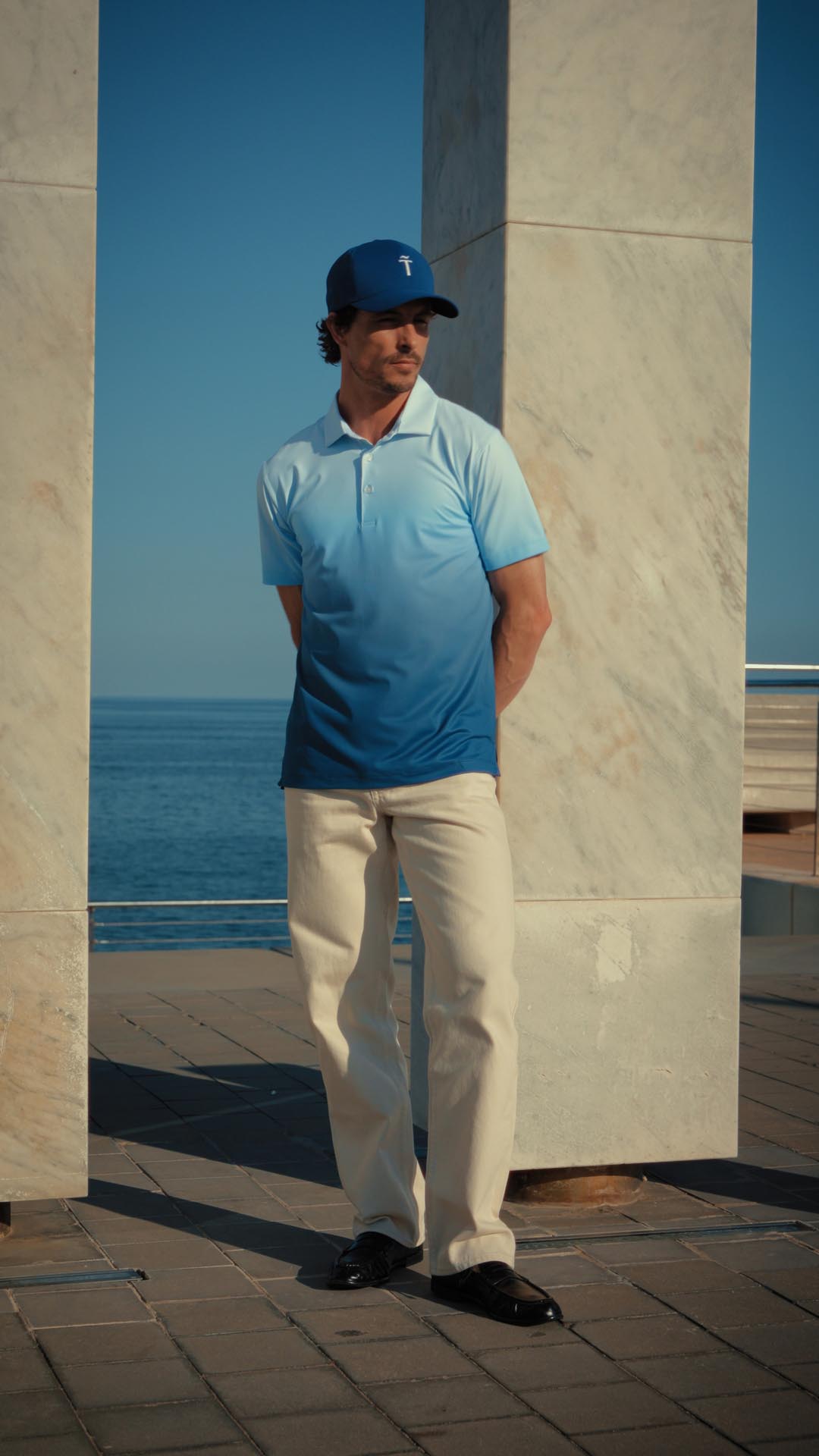 Man wearing a blue polo shirt and cap standing by a marble column with a clear blue sky and ocean in the background.