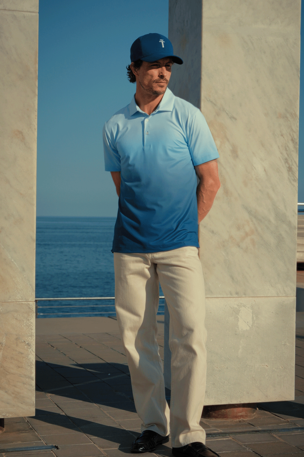 Man wearing a blue polo shirt and cap standing by a stone wall with a clear blue sky and ocean in the background.