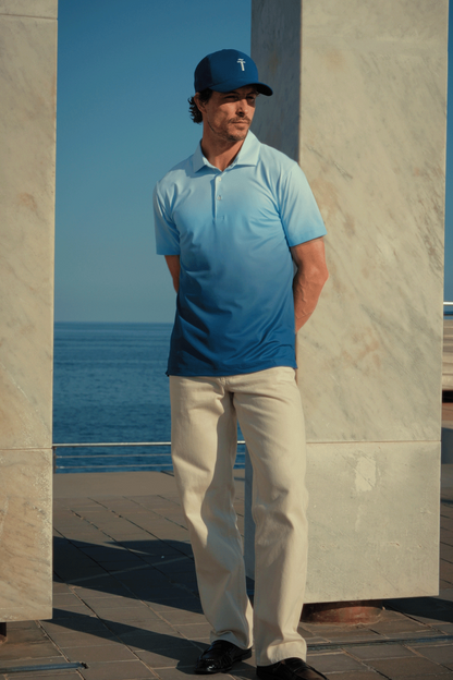 Man wearing a blue polo shirt and cap standing by a stone wall with a clear blue sky and ocean in the background.