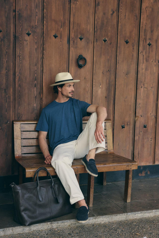 Man sitting on a wooden bench wearing a blue shirt, white pants, and a Panama hat, with a black bag beside him against a wooden wall.