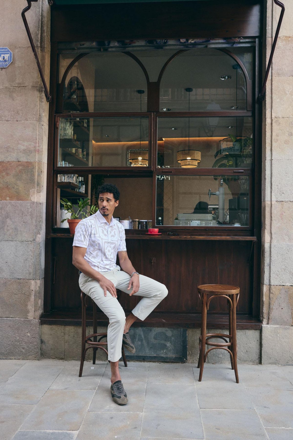 Man sitting on a stool outside a cafe with stone walls and glass windows.
