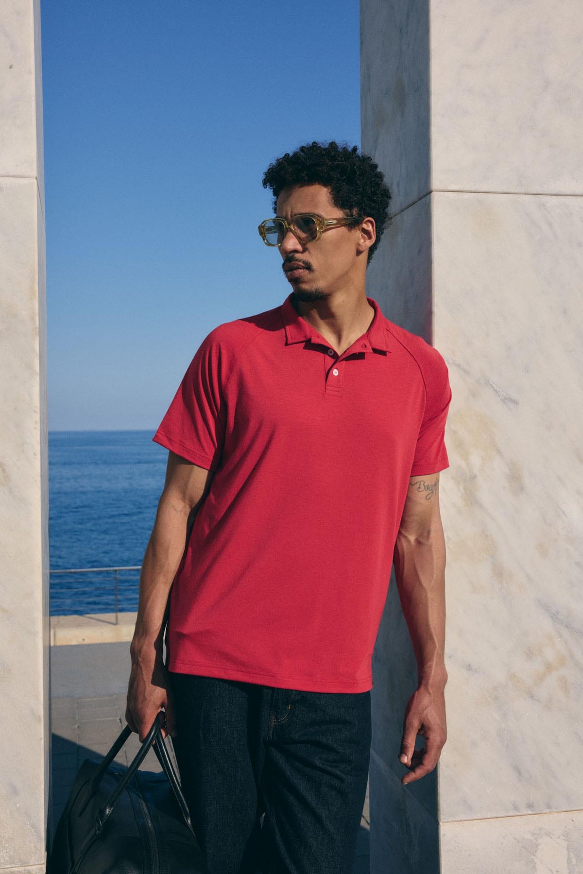 Man wearing a red polo shirt standing by a marble wall with a blue sky and ocean view.
