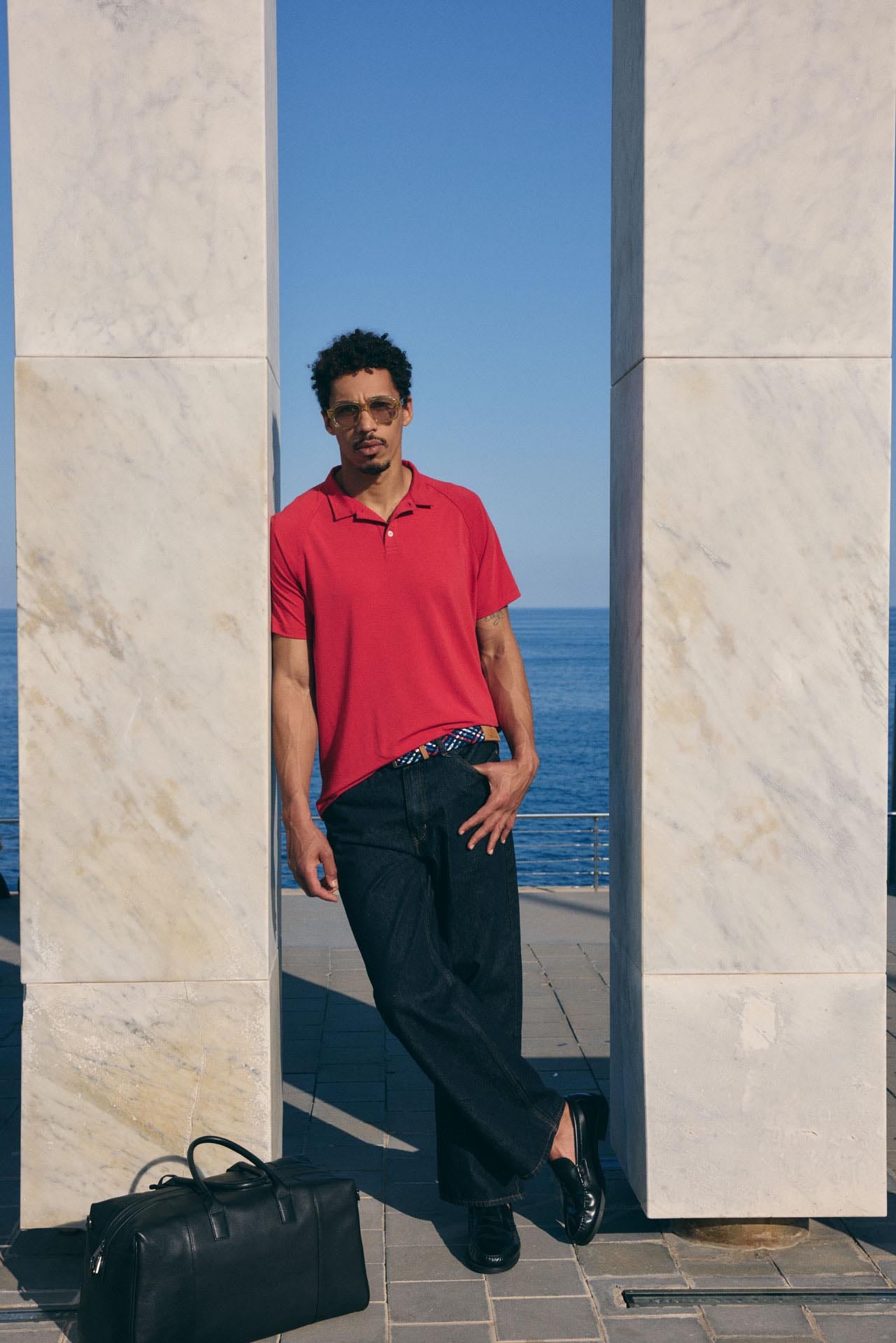 Man in a red shirt standing by a stone wall with a blue sky and water in the background