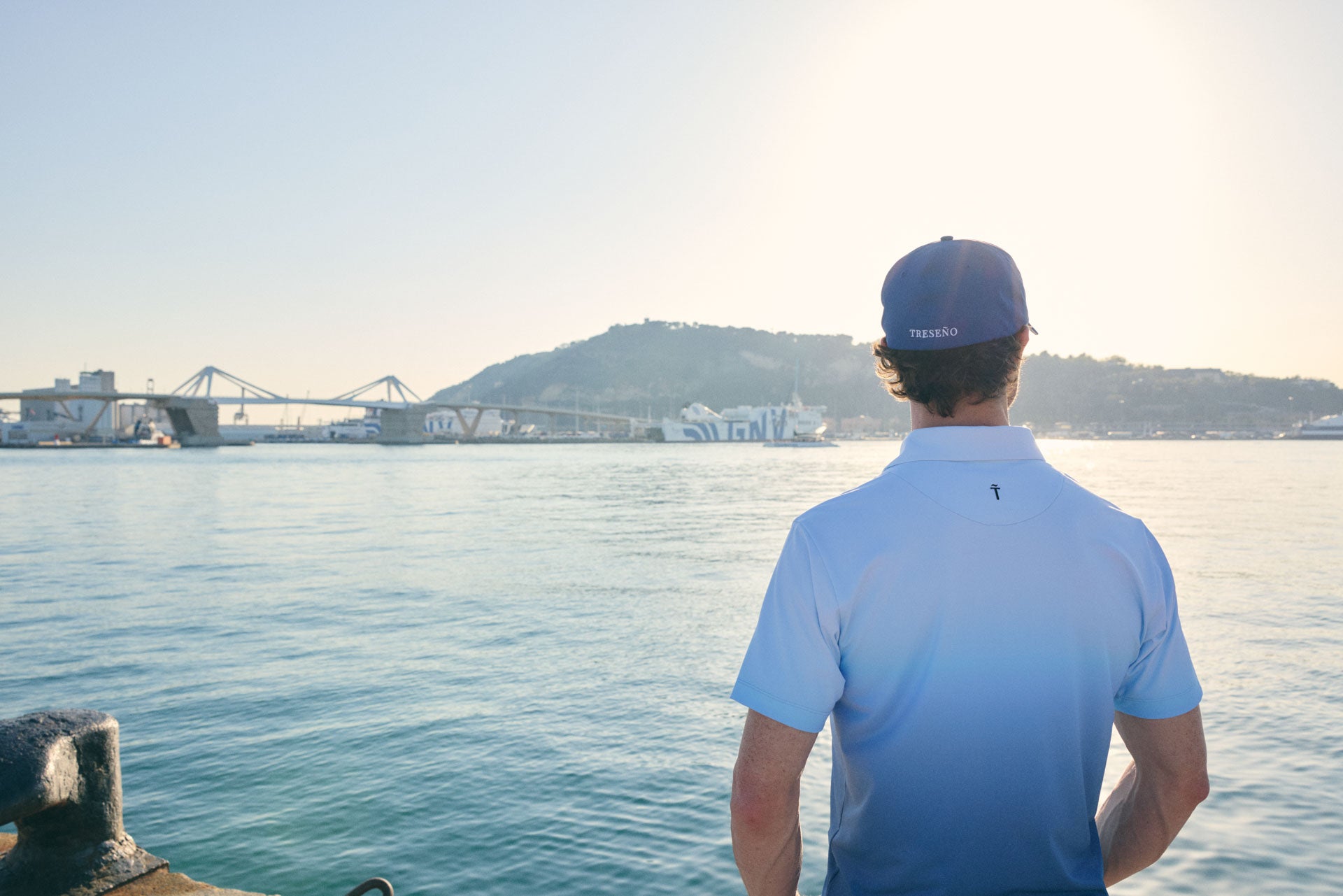 Man wearing a blue cap and light blue shirt standing by a waterfront with a bridge and hills in the background.