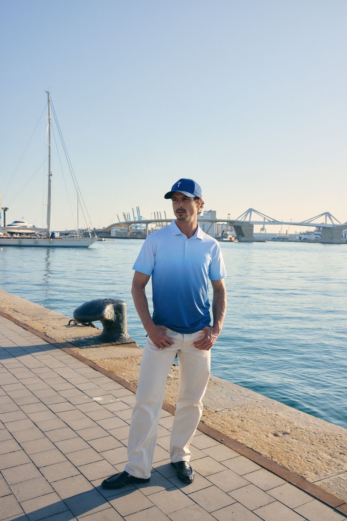 Man standing by a waterfront with a sailboat and bridge in the background