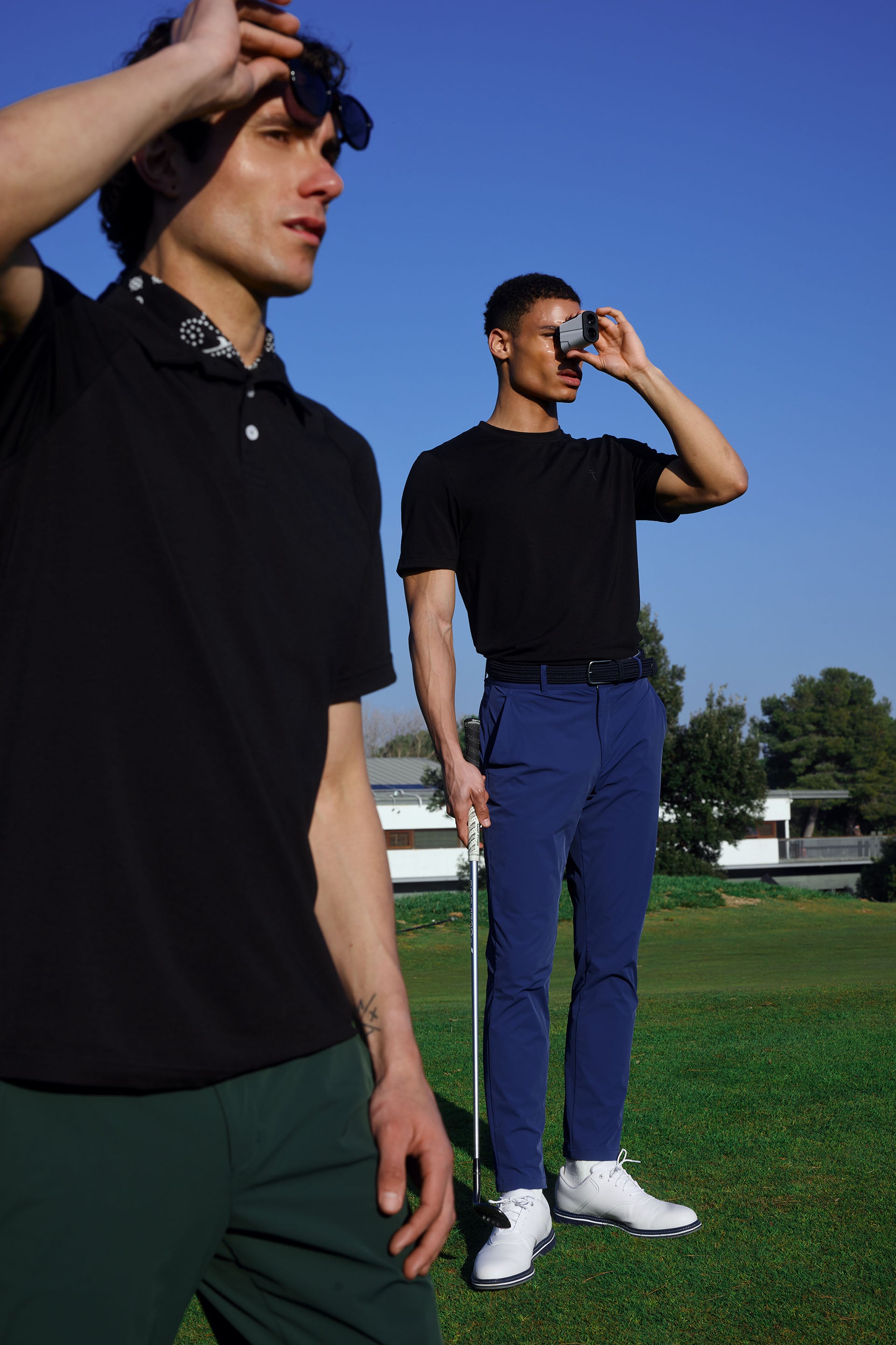 Two men on a golf course with one adjusting his sunglasses and the other holding a golf club.