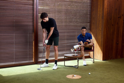 Two men playing practicing putting with a wooden wall and green floor.