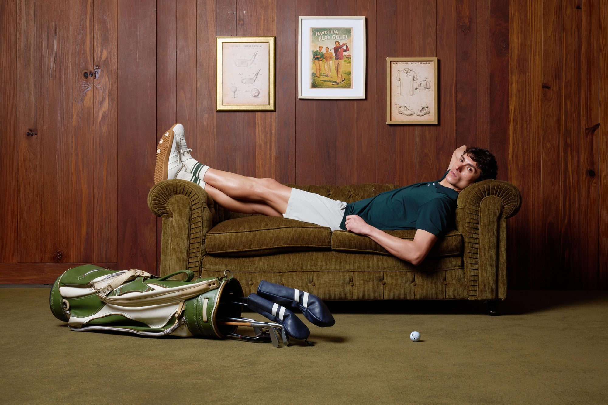 Man golfer lying on a brown couch in a room with wooden walls and sports equipment wearing Treseño golf clothing