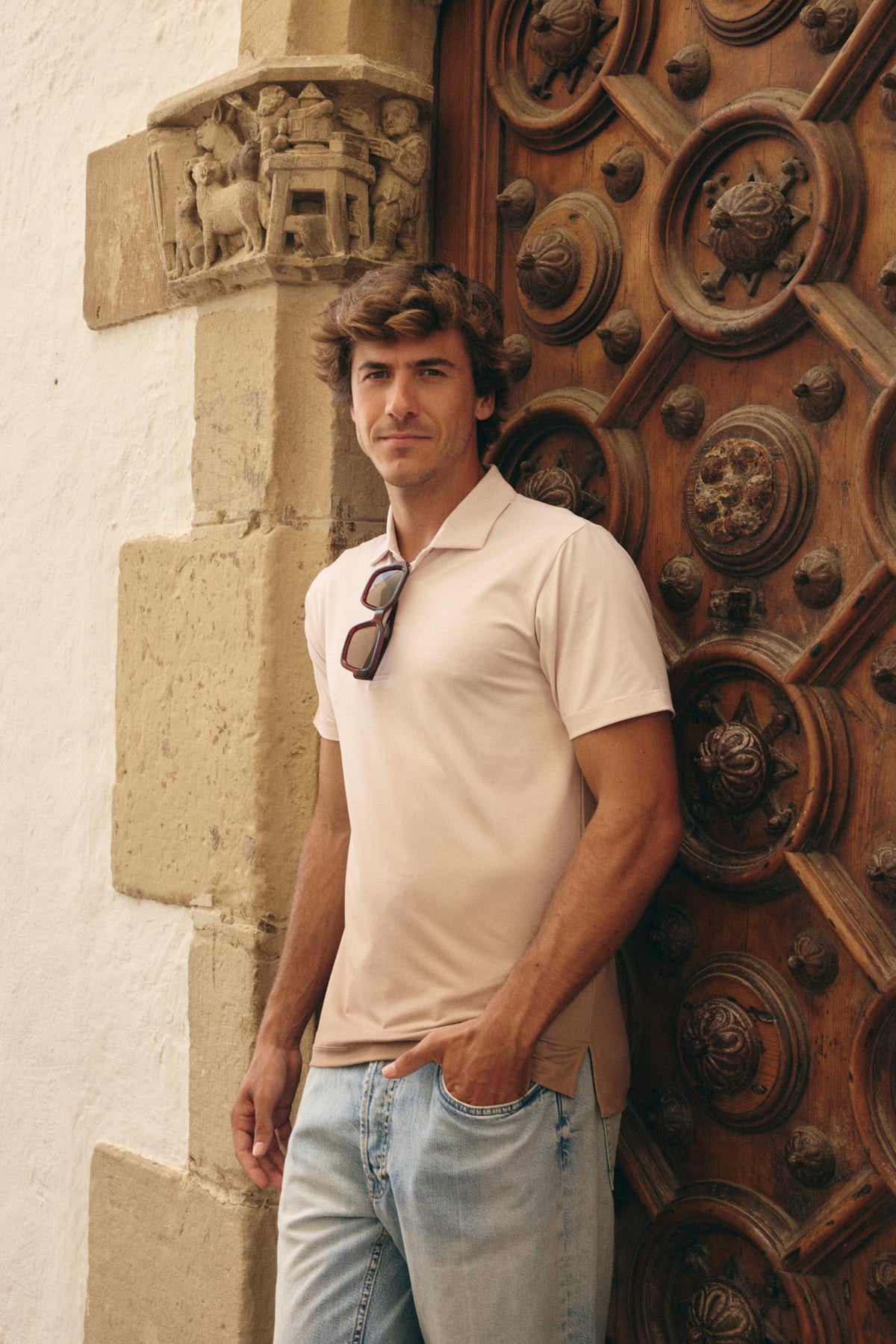 A man wearing a light beige short-sleeve polo shirt and light blue jeans stands leaning against a large ornate wooden door next to a white stucco wall.
