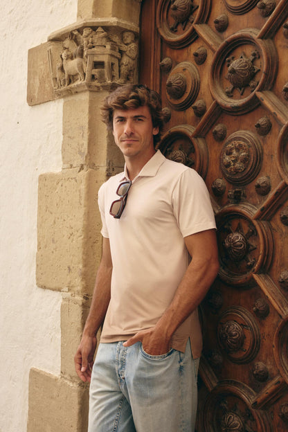 A man wearing a light beige short-sleeve polo shirt and light blue jeans stands leaning against a large ornate wooden door next to a white stucco wall.