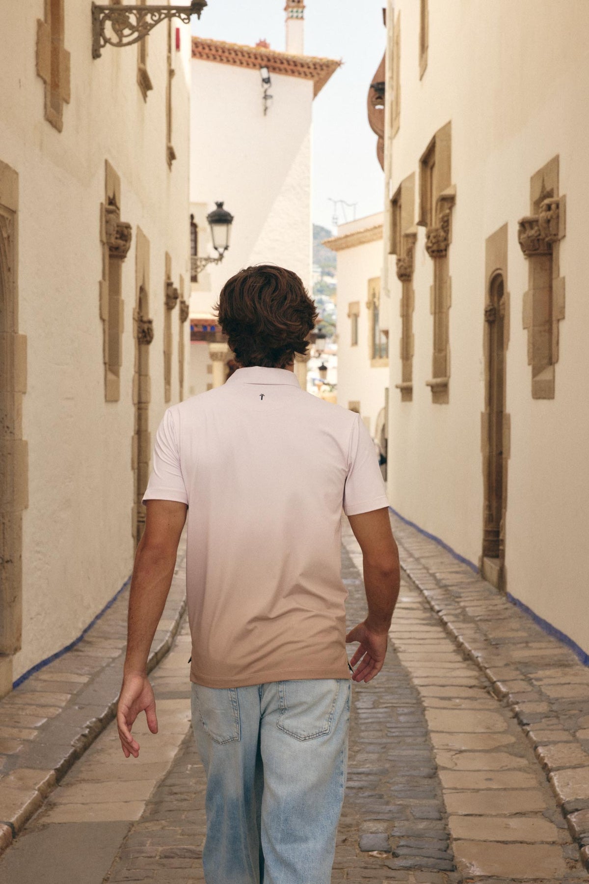 A man viewed from behind walking down a narrow, sunlit cobblestone alleyway lined with warm beige stucco buildings. He is wearing a light beige and white gradient short-sleeve polo shirt and light blue jeans.