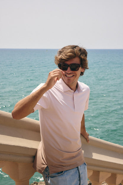A smiling man wearing a light pink polo shirt and sunglasses, leaning on a stone railing with the blue ocean in the background.